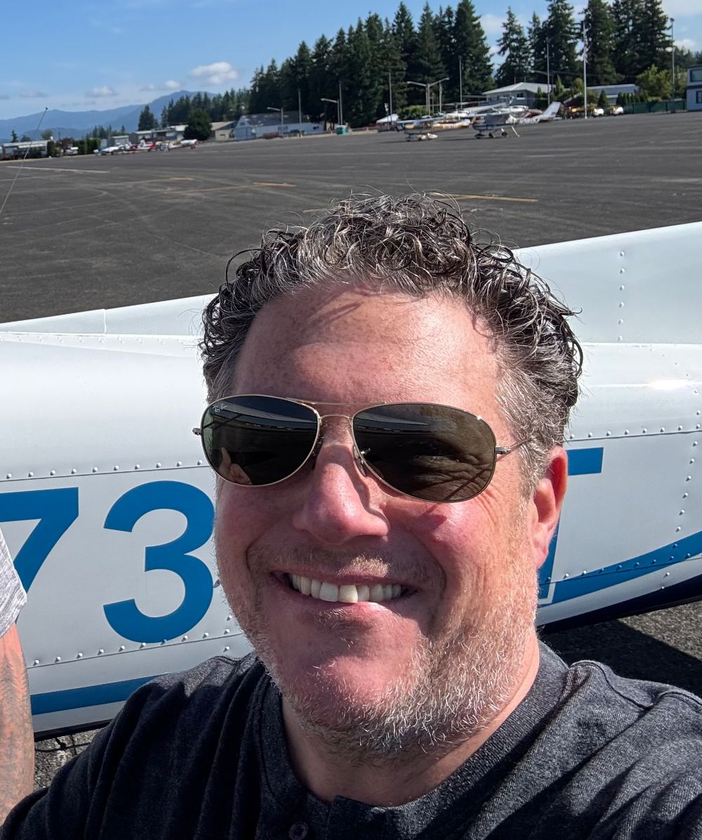 Steve Kann in sunglasses, smiling, beside a light airplane on the ramp.