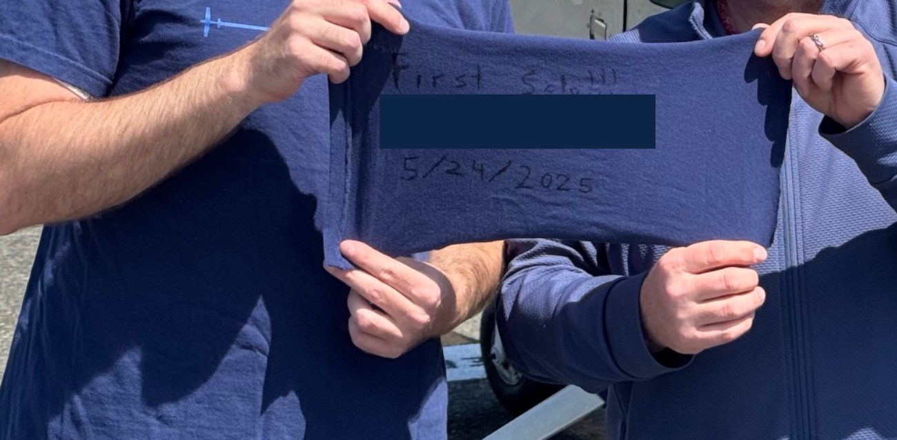Two people holding a navy first-solo cloth dated 5/24/2025. The name on the cloth is covered.